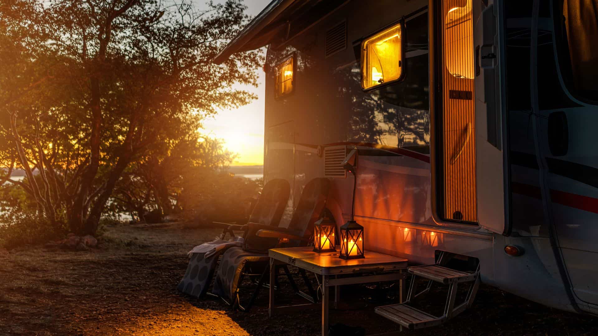 A parked RV at sunset with two chairs and a table holding lanterns outside, nestled among trees by a peaceful lake—an inviting scene perfect for campground marketing.