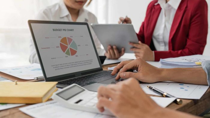 Three people sit at a desk reviewing documents; one uses a laptop displaying a budget pie chart, another from an SEO company in Toronto holds a calculator, while the third works on a tablet.