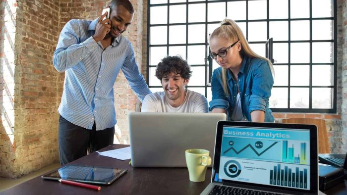 Three people work together at a desk with laptops and documents as business analytics data, relevant for an SEO company in Toronto, is displayed on a screen in the foreground.