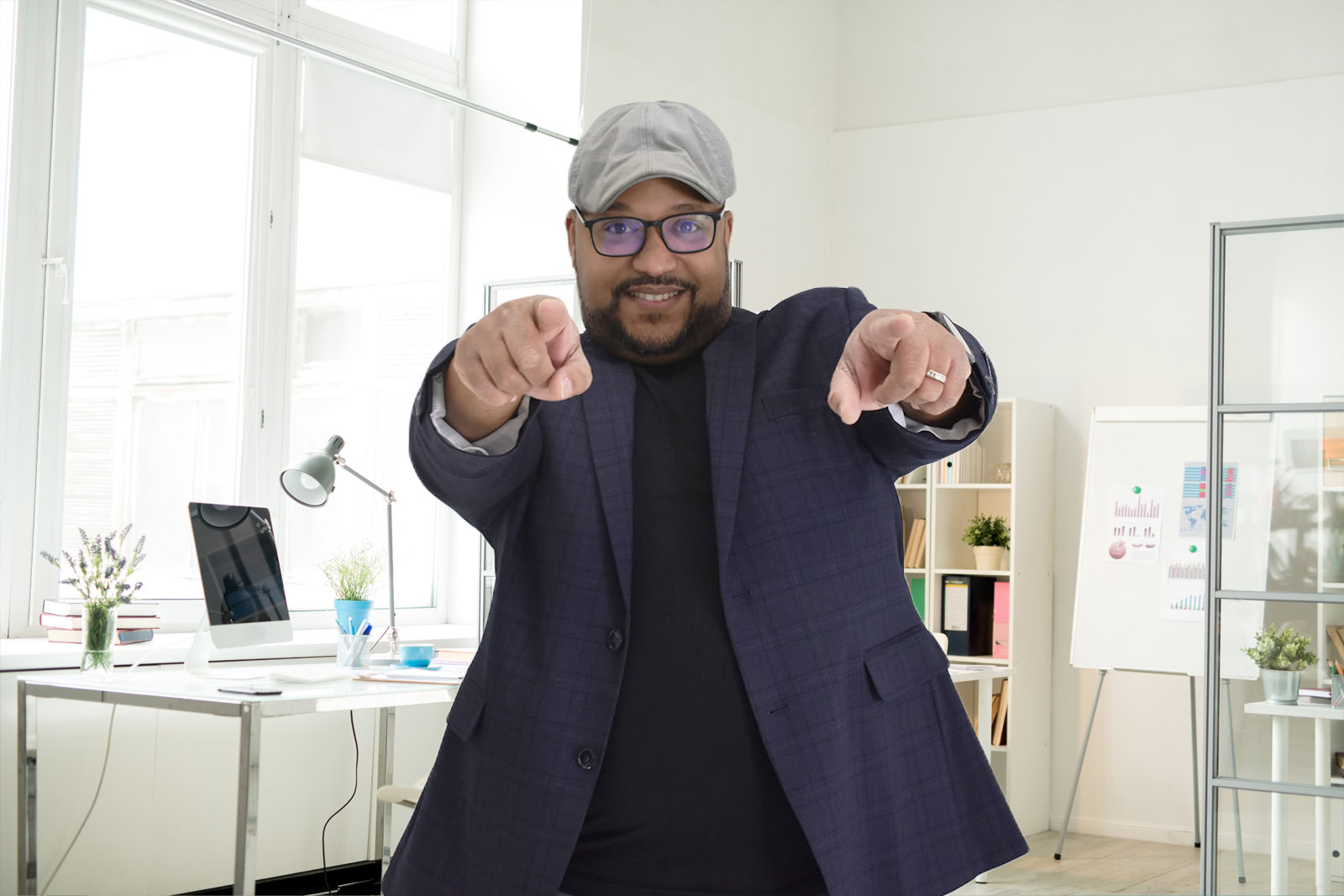 A man wearing glasses, a cap, and a blazer stands in an office space, smiling and pointing towards the camera with both hands, embodying the confidence of a professional offering digital marketing service.