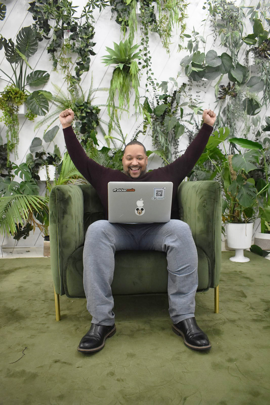A man sits on a green armchair with his arms raised in excitement while using a laptop, embodying the enthusiasm of digital marketing experts, surrounded by potted plants and lush greenery.
