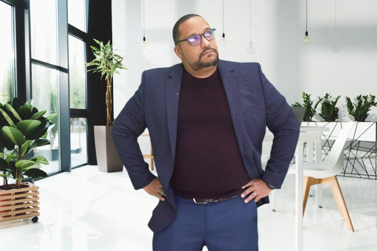 A man in a blue suit and glasses stands with hands on hips in a modern, bright office with plants and white furniture, ready to discuss local SEO services.