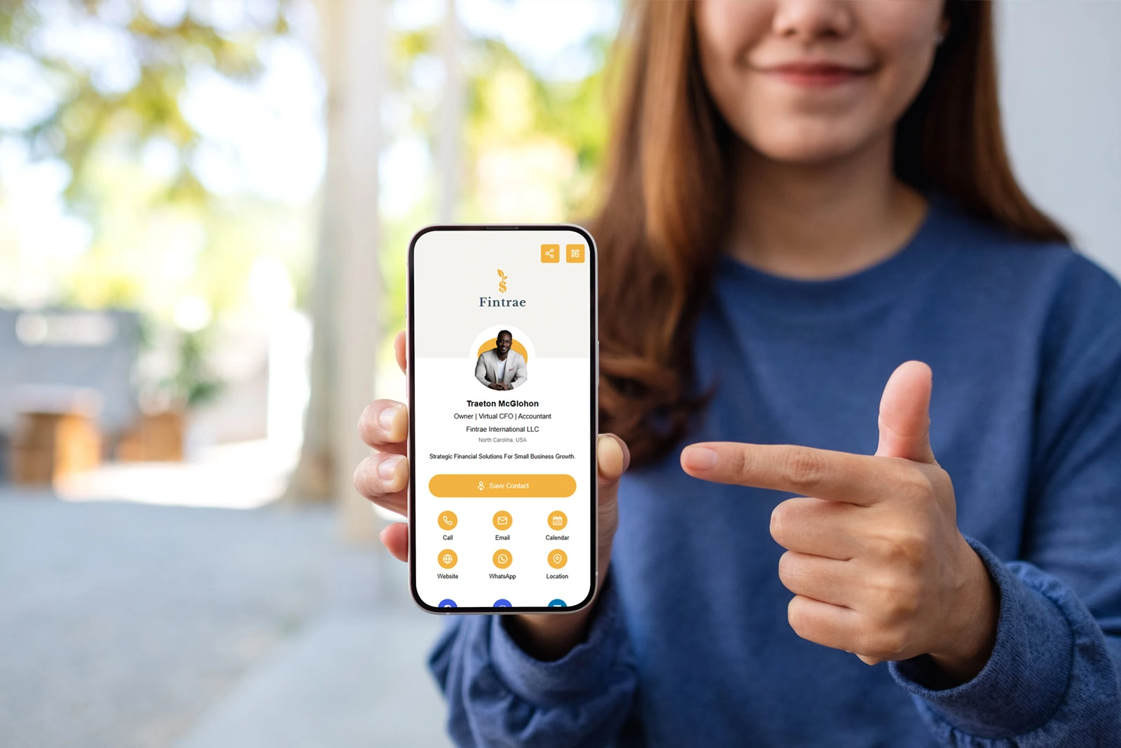 A woman in a blue sweater holds a smartphone displaying a digital business card on a business networking app, pointing at the screen.